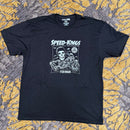 A black short-sleeve graphic t-shirt featuring a white and gray 'Speed-Kings' motorcycle print, laid flat on a textured yellow and gray patterned carpet.