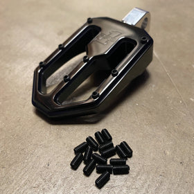 Black flat bicycle pedal with pile of black set screws (replacement pins) on concrete surface